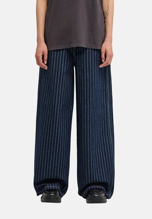 Personne portant un pantalon large à rayures verticales bleu foncé, des chaussures noires et une chemise gris foncé, debout avec les mains détendues le long du corps.