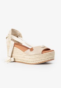 Espadrille de cuña con parte superior de tela beige texturizada, plantilla color marrón y suela de yute trenzado. Presenta cintas de adorno y un detalle decorativo.