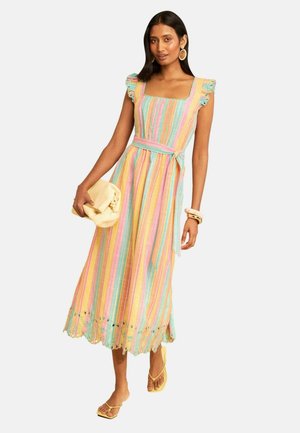 Kvinde iført en flerfarvet, stribet ærmeløs kjole med flæseærmer, der holder en beige clutch og har gule sandaler og armbånd på.