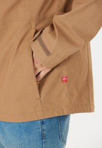 Beige jacka i lätt material, med en sidficka, kontrasterande sömmar och en liten röd och vit flaggdetalj.