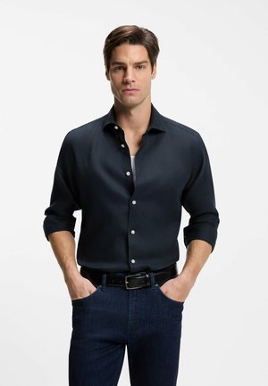 Mann mit Händen in den Taschen, trägt ein dunkelblaues Hemd mit Knöpfen, hochgekrempelte Ärmel und dunkle Jeans vor einem schlichten weißen Hintergrund.