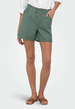 Femme portant un short vert à taille élastique et des sandales marron à enfiler, debout avec une main dans la poche.