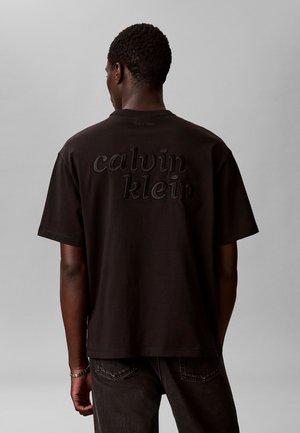 Man draagt een zwarte Calvin Klein T-shirt met groot reliëflogo op de achterkant, van achteren getoond tegen een effen grijze achtergrond.