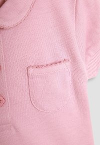 Puha rózsaszín kötött anyag kis hullámos szegélyű zsebbel, valamint részleges kilátás egy gombra és gallérra egy ruhadarabon.