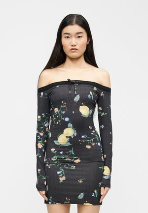 Noor naine, kellel on pikad tumedad juuksed, kandes kehakinnitusega, õlgadest paljast musta kleiti sidruni- ja lillemustriga, seismas ühevärvilise tausta ees.