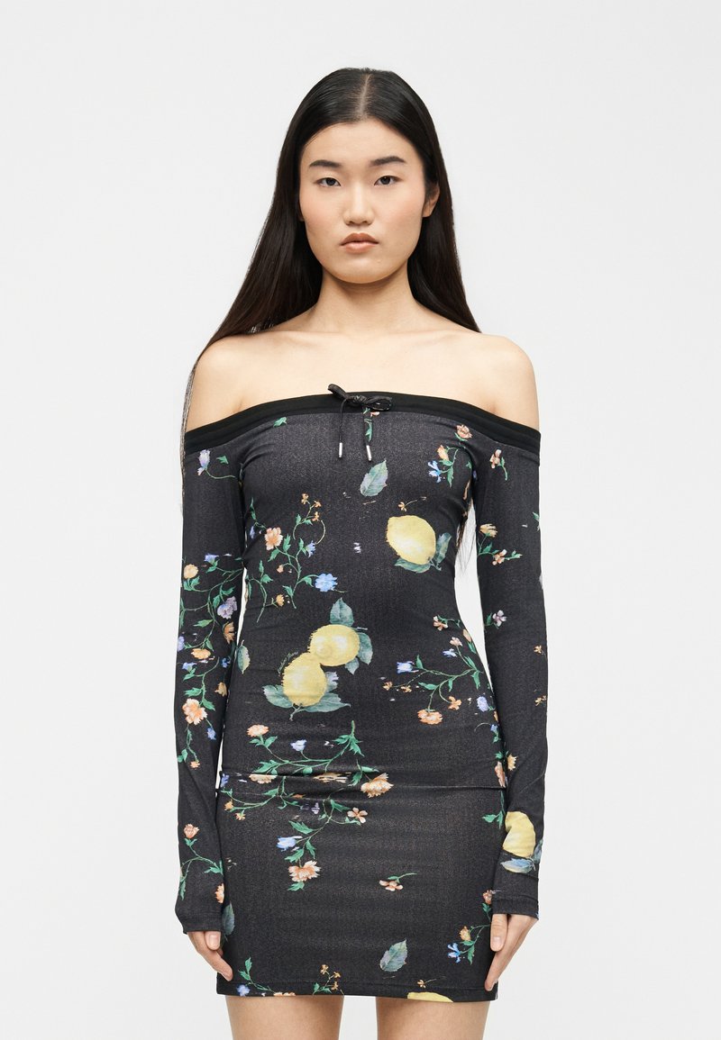 Νεαρή γυναίκα με μακριά σκούρα μαλλιά, φοράει εφαρμοστή μαύρη φόρεμα με ανοιχτούς ώμους, διακοσμημένη με λεμονί και ανθικό σχέδιο, στέκεται μπροστά σε απλό φόντο.