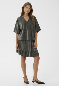 Woman standing wearing a loose dark gray glittery V-neck top with wide sleeves and matching ruffled skirt, black pointed slingback heels.