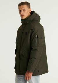 Chaqueta de invierno verde oliva con capucha, detalles de parches y bolsillos con cremallera. Fabricada con material impermeable, cuenta con una textura acolchada y costuras resistentes.