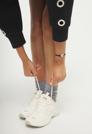 Mains nouant des baskets blanches avec des chaussettes grises, portant un pantalon noir avec de grands œillets argentés et un bracelet noir avec un pendentif cœur argenté.