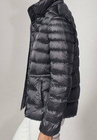 Sort dunjakke med quiltet design, høj krave og sidelommer. Lavet af glat, skinnende stof med ribbet detaljer.