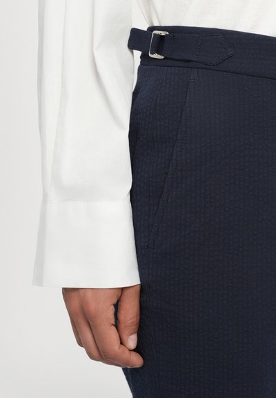 Pantalon à motifs navy avec une finition texturée, présentant une ceinture avec une boucle en argent et des poches latérales. Détail d'une chemise blanche à manches longues visible.