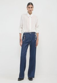 Donna che indossa una camicia bianca trasparente a righe con bottoni, jeans a vita alta e a gamba larga di colore blu scuro con bottoni laterali, e scarpe nere.