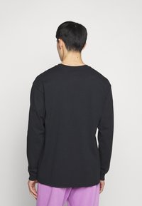 Svart långärmad T-shirt med rund halsringning, i avslappnad passform med ribbade ärmar, sedd från baksidan, matchad med lila byxor.