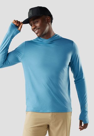 Homme souriant portant une chemise bleu vif à manches longues avec des trous pour le pouce et un pantalon beige, ajustant une casquette de baseball noire.