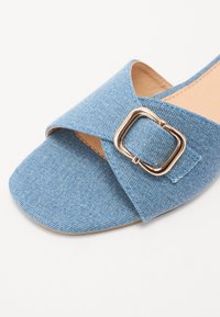 Denim sandaler med korskonstruktion, ljusblå färg, en fyrkantig guldspänne som accent, och en platt sula. Slät textur.