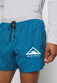Gros plan d'une personne portant un short Nike Trail bleu avec des lignes topographiques et un cordon noir, associé à un haut à manches longues gris clair.