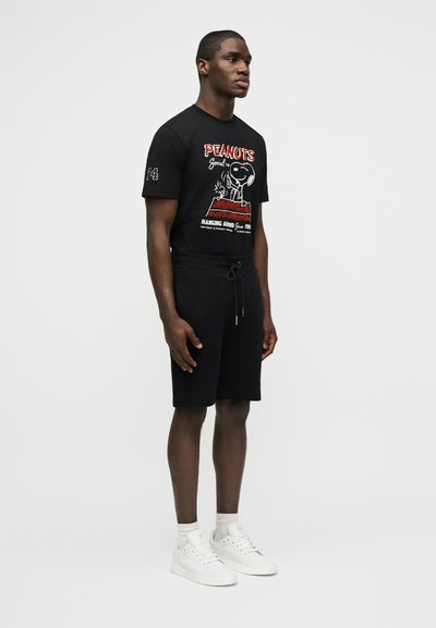 Homme debout portant un T-shirt noir avec un motif Peanuts, un short noir avec cordon de serrage, des chaussettes blanches et des baskets blanches, devant un fond uni.