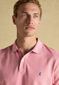 Gaiši rozā kokvilnas polo krekls ar klasisku apkakli, uz krūtīm ar zilu izšūtu logo un divu pogu aizdari.