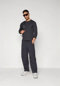 Black ribbed long-sleeve shirt, dark gray cargo pants with pockets, and light gray sneakers. Sunglasses and a backpack complete the outfit.