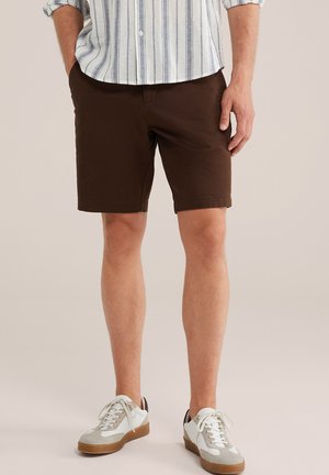 Mand iført mørkebrune shorts, hvide og grå sneakers og en lys skjorte med opkædede ærmer, der står mod en ensfarvet baggrund.