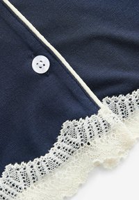Marineblauer Strickstoff mit einem weißen Spitzenbesatz am unteren Rand. Verfügt über einen runden weißen Knopf und eine kontrastierende weiße Paspelierung entlang der Vorderseite.