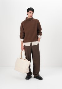 Καφέ ριγωτό πουλόβερ με ψηλό λαιμό, φορεμένο πάνω από ένα ανοιχτό πουκάμισο. Καφέ παντελόνι, μαύρες σαγιονάρες και μία μπεζ καμβά τσάντα. Λευκό φόντο.