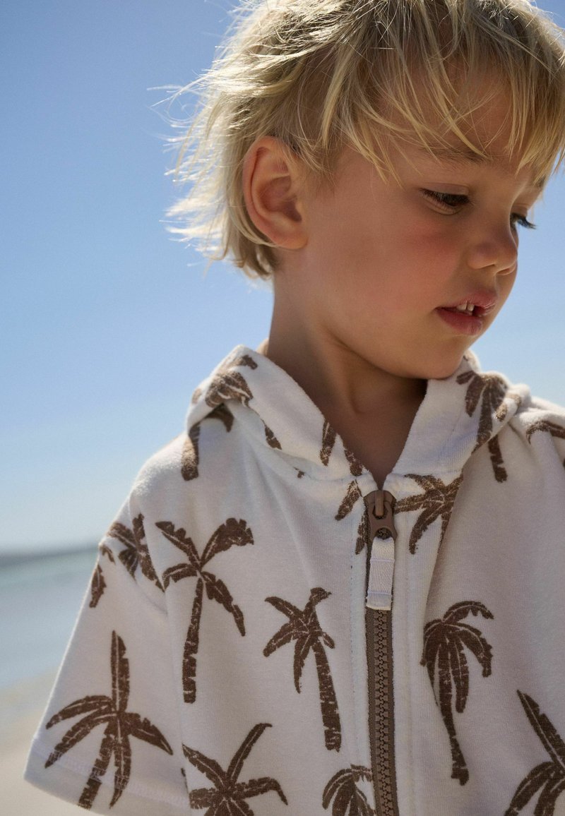 Mladé dieťa s blond vlasmi, oblečené v bielej mikine s hnedými potlačami palmových stromov, pozerajúce sa dolu na pozadí jasnej modrej oblohy.