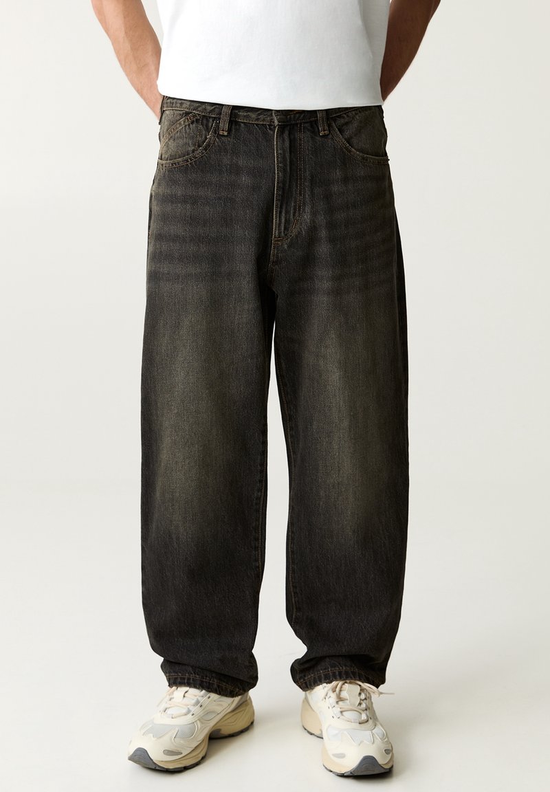 Personne portant un jean noir délavé à coupe ample et des baskets blanches, debout les mains dans les poches devant un fond uni.