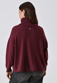Bordeauxfarbener Pullover mit hohem geripptem Kragen und langen Ärmeln. Mit einem Logodetail auf der Rückseite und einer lockeren Passform. Weicher, strukturierter Stoff.