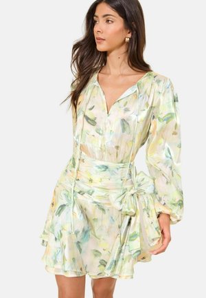 Kobieta nosząca pastelową, kwiatową sukienkę z długim rękawem, z głębokim dekoltem w serek i szerokim pasem w talii zawiązanym na kokardę po prawej stronie.