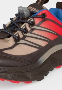 Chaussure de trail running beige, rouge et marron, col matelassé bleu et lacets élastiques bleu et noir à motifs maintenus par un bouton de serrage gris.