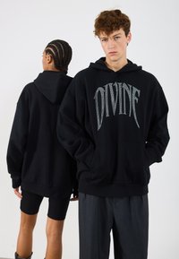 Hanorac negru oversized cu un grafic argintiu "DIVINE", având un buzunar tip cangur și glugă cu șnur, purtat de doi modele.