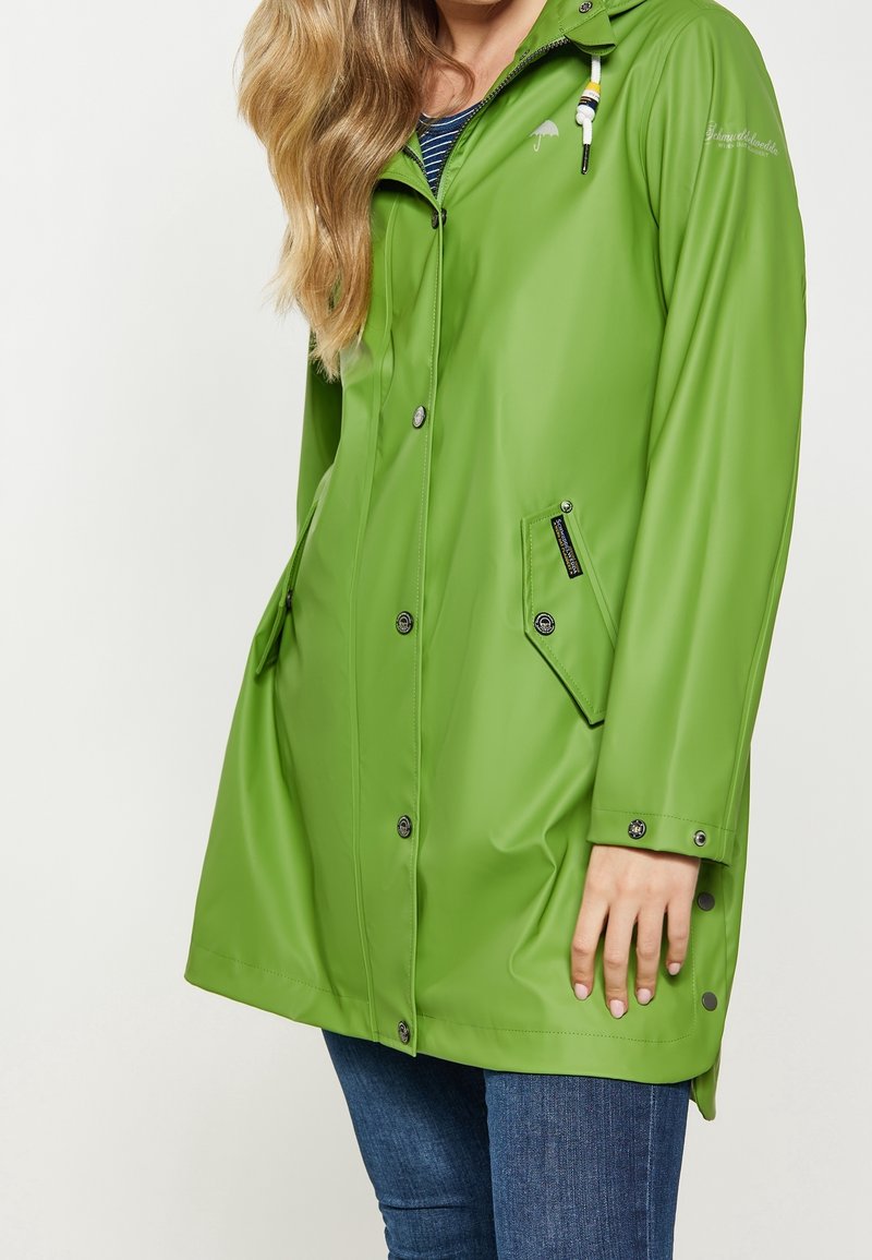 Femme portant un long imperméable vert vif avec des boutons-pression à l'avant et des poches à rabat sur un jean bleu, debout devant un fond blanc uni.
