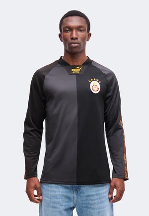 Homme portant un maillot de football Puma noir bicolore à manches longues avec le logo Galatasaray et une bande à motifs sur les manches, assorti à un jean bleu.
