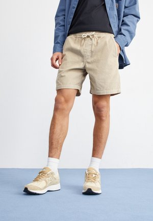 Shorts en velours côtelé beige avec une taille élastique et un cordon de serrage, portés avec une chemise bleu marine et des baskets beige à semelles blanches et des chaussettes claires.