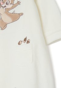 Monos de tela color crema con un personaje bordado en la parte delantera y un bolsillo con diseños de huellas de patas en bordado marrón.