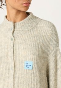 Svetlo bež pletena jopica z rebrasto zasnovo, s visokim ovratnikom, z gumbi in modro oznako z napisom "LA MAISON AMV."