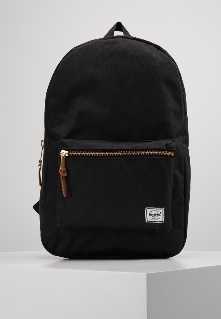 Sac à dos en tissu noir avec une poche zippée à l'avant, des tirettes de fermeture éclair marron et une petite étiquette de marque blanche rectangulaire dans le coin inférieur droit.