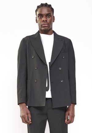 Man met gevlochten haar draagt een zwarte dubbelgeknoopte blazer over een wit overhemd, staand tegen een effen witte achtergrond.