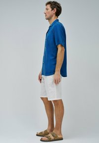 Camisa de manga corta y botones en tejido azul, combinada con pantalones cortos blancos y sandalias planas marrones. Textura suave con un ajuste relajado.