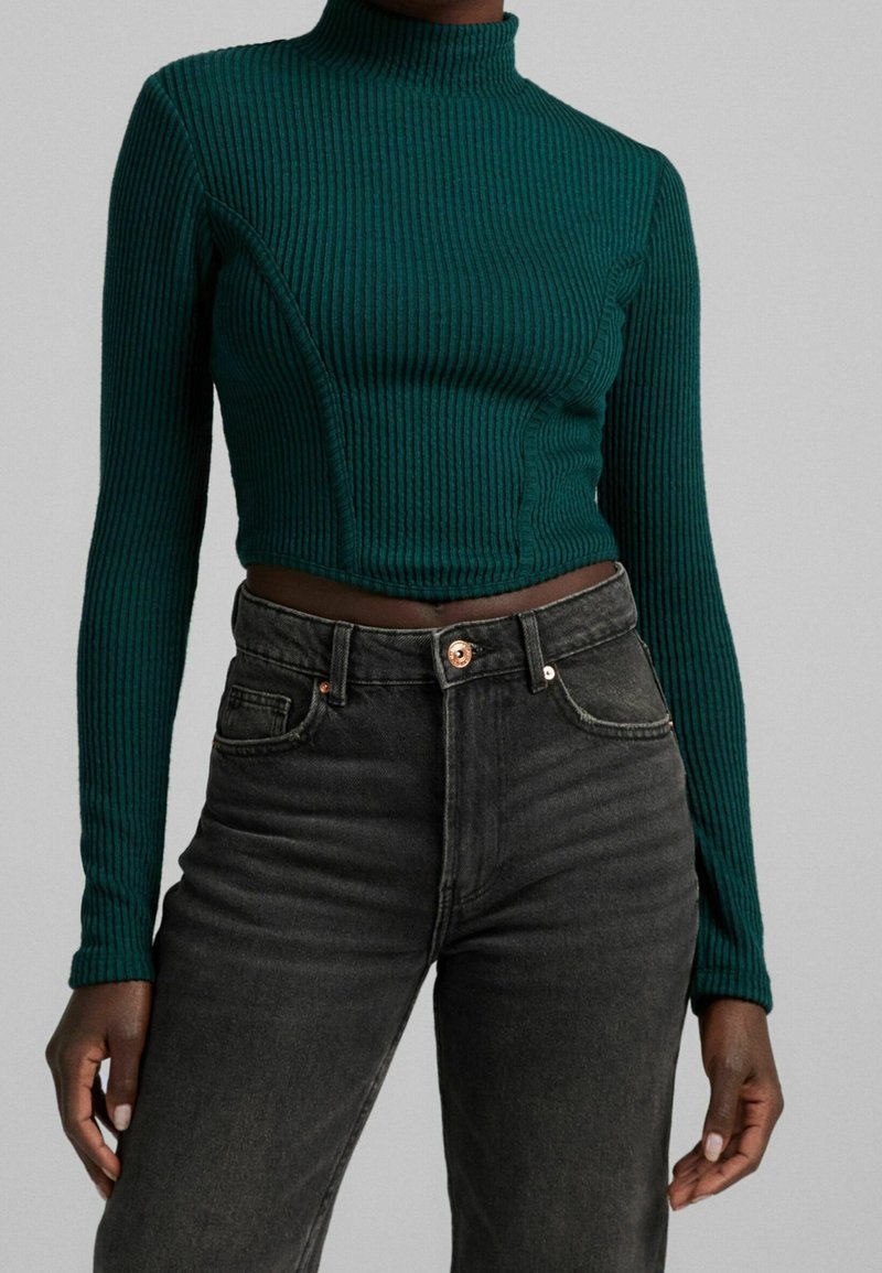 Top a collo alto a coste verde smeraldo con maniche lunghe e un design corto aderente, abbinato a jeans scuri a vita alta.