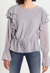 Blusa grigio chiaro con maniche a ruffle sovrapposte, vita elastica e dettaglio con cordoncino. Il tessuto semi-trasparente e le maniche a campana aggiungono texture.