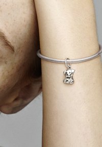 Pulsera de plata con una textura trenzada, que presenta un pequeño colgante de perro con detalles finos y un acabado brillante, reposando sobre un tono de piel clara.
