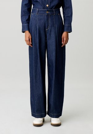 Personne portant un pantalon en denim bleu foncé taille haute à jambes larges et une chemise en denim assortie, associée à des chaussures blanches sur un fond uni.
