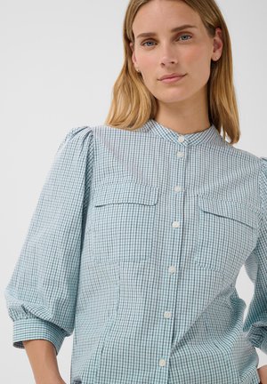 Jeune femme aux cheveux blonds portant une chemise à carreaux bleu clair avec des manches bouffantes et deux poches poitrine, debout devant un fond uni.