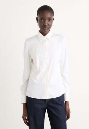Una camicia bianca con colletto a punta, maniche lunghe e spalle leggermente arricciate, abbinata a jeans di denim blu scuro.