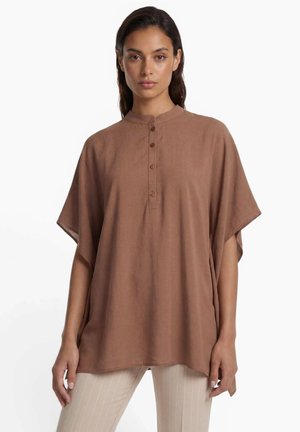 Jonge vrouw draagt een losse bruine top met knopen en korte mouwen en lichtbeige broek met fijne strepen tegen een witte achtergrond.