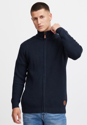 Suéter de punto azul marino con cremallera, con cuello y puños acanalados, que presenta un acento de cuero en el dobladillo y un patrón texturizado en todo.