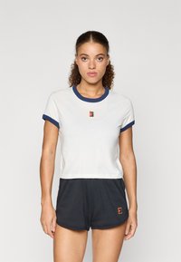 Λευκό κοντό μπλουζάκι με ναυτικό μπλε ριγωτό γιακά και μανίκια; διαθέτει ένα μικρό, πολύχρωμο λογότυπο στο στήθος. Συνδυάζεται με σκούρες βερμούδες.