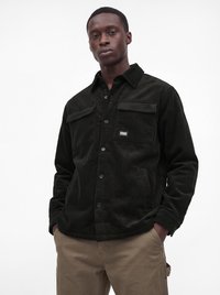 Man wearing a black corduroy button-up jacket with front pockets and beige pants, standing with hands in pockets against white background.